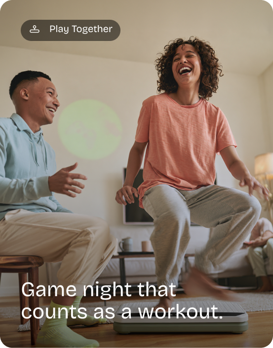 Woman laughs mid-squat on the Kiwii Move board while a friend cheers from a stool. Play Together: Game night that counts as a workout.